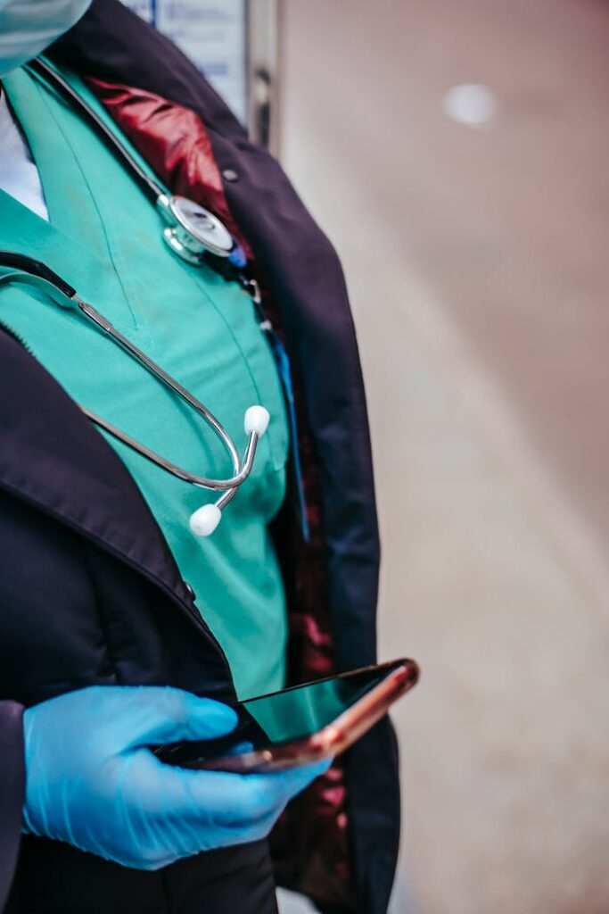 Crop anonymous female doctor in uniform with stethoscope touching black screen on cellphone in town